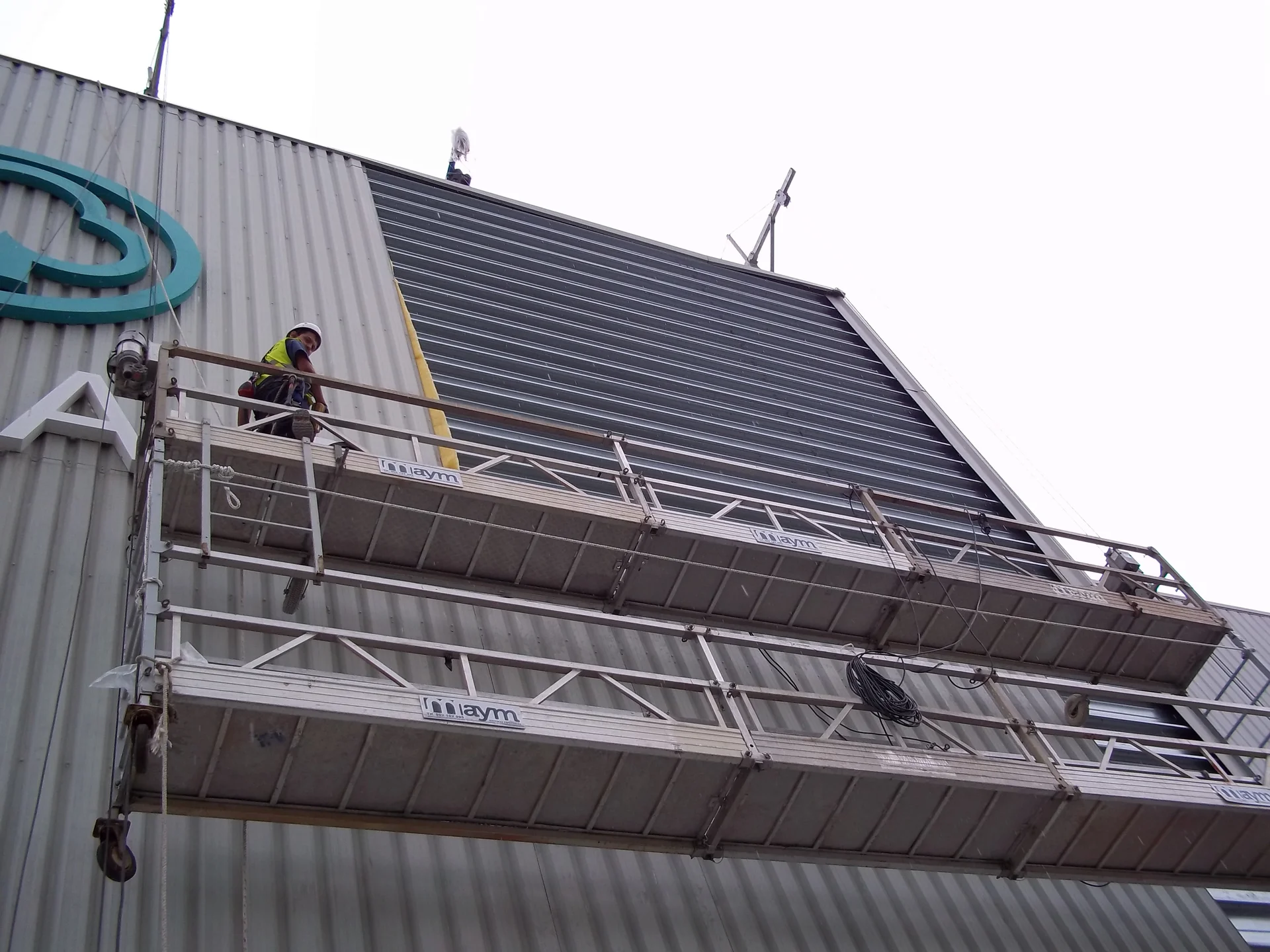 Operario trabajando con andamio eléctrico suspendido en fachada metálica de nave industrial, empresa asociada a ALEA